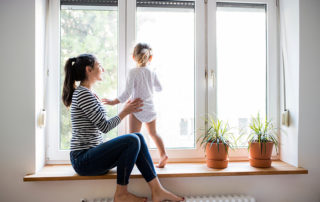 A mom holding a child on a thick window sill as she looks out the windows