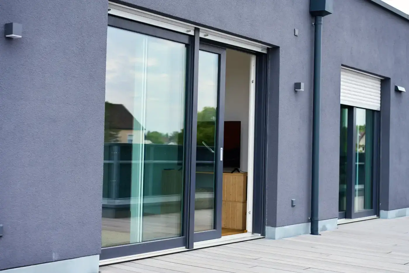 High-Quality Sliding Patio Doors In Minnetonka, MN | Zen Windows Twin Cities The back wall of a multi-home building, showing a dark blue exterior, a gray wooden deck, and sliding patio doors with dark blue framing. The door closest to the camera is partially opened, and through the glass, you can see a wooden TV stand and a flat-screen TV on top of it.